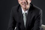 A person in formal attire, wearing a suit and tie, is seated and smiling against a dark background.