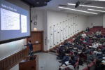 A person presenting a lecture in a classroom with a projected slide titled "ResNets in PyTorch: a Noted." Students are seated in tiered rows, attentively listening.