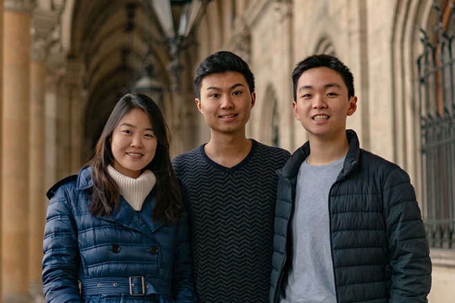 Three people standing closely together, smiling, in an outdoor setting with architectural columns in the background.