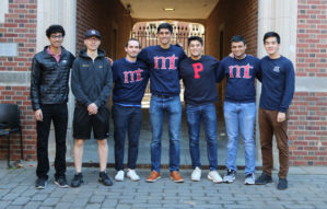 A group of six people standing in a row outside, in front of a brick building. Some are wearing sweatshirts with letters on them. They are casually dressed and smiling.