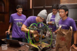 A group of people in purple PennApps XV shirts working on a tech project involving wires and tubes, with a laptop nearby.