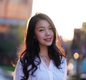 Portrait of a person with long dark hair and a white shirt smiling softly, with a softly blurred urban background and warm backlighting.