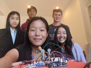 A group of five smiling people, two in front and three at the back, are gathered around an electronic project with wires on a table.