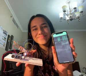 A person holding a breadboard with electronic components and a smartphone displaying a graph, possibly showcasing a tech project or experiment.