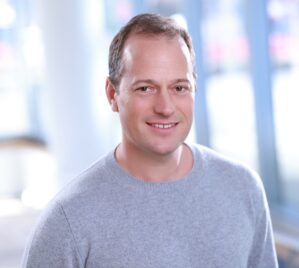 A headshot of a person wearing a light gray sweater, smiling, with a blurred background.