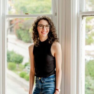 Person with curly hair and glasses stands by a window, wearing a sleeveless black top and jeans, smiling. Bright, natural light fills the room.