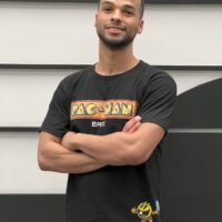 A person stands with crossed arms, wearing a black Pac-Man t-shirt and smiling, in front of a minimalist background with horizontal lines.