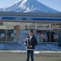 A person in winter clothing stands in front of a convenience store, with Mount Fuji in the background under a clear blue sky.