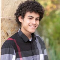 Smiling individual with curly hair wearing a plaid shirt, leaning against a stone wall with a blurred green background.