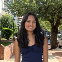 A person is smiling while standing on a sidewalk with trees and buildings in the background.