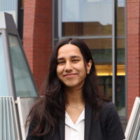 A person with long dark hair smiling, wearing a black blazer and white shirt, stands in front of a building with triangular glass structures.