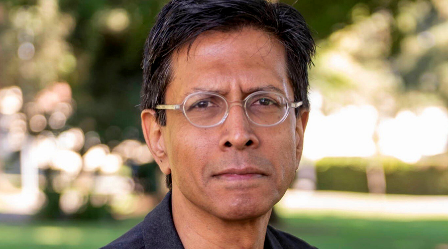 Headshot of a person with short, dark hair, wearing glasses and a dark jacket, standing outdoors with a blurred background of trees and greenery.