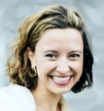 Headshot of a person with short, light brown hair, smiling, wearing hoop earrings. The background is softly blurred.