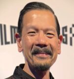 This image is a headshot of a person with short, slicked-back hair and a mustache, smiling slightly. The background appears to be an event or exhibition with indistinct text visible.