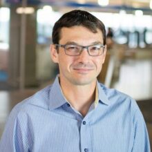David Quattrone A headshot of a person wearing glasses and a blue striped shirt. The background is blurred, suggesting an indoor office environment.