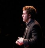 A person in profile view giving a talk or presentation on stage, wearing a dark suit.