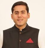 Headshot of a person wearing a black Nehru jacket with a red pocket square, over a white shirt, against a plain background.