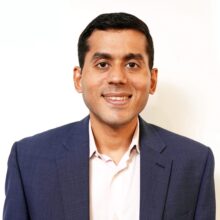 Nalin Moniz - Photo High Resolution Headshot of a person wearing a blue suit jacket and light-colored shirt, smiling, with a plain white background.