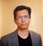 Person wearing glasses, a black shirt, and a gray blazer, looking at the camera against a neutral background. Headshot style image.