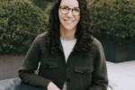 A headshot of a person with curly hair and glasses, sitting at an outdoor table, wearing a dark jacket. Bushes are in the background.