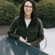 Unknown A headshot of a person with curly hair and glasses, sitting at an outdoor table, wearing a dark jacket. Bushes are in the background.