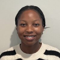 professhy headshot Headshot of a person with braided hair, wearing a black and white striped sweater, smiling against a neutral background.