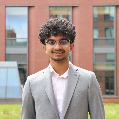 Headshot of person wearing glasses and a suit, standing in front of a brick building with large windows.