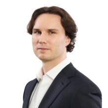 Headshot of a person with medium-length dark hair, wearing a dark blazer over a white shirt, set against a plain background.