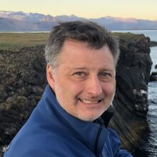 Headshot of a person outdoors with rocky cliffs and water in the background. Snow-capped mountains are visible in the distance.