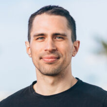 Headshot of person with short dark hair, wearing a black shirt, standing outdoors.