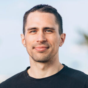 Headshot of person with short dark hair, wearing a black shirt, standing outdoors.